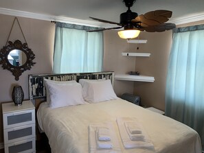 Een net opgemaakt bed met witte lakens en handdoeken, een nachtkastje met een lamp, een plafondventilator en een plank met boeken.