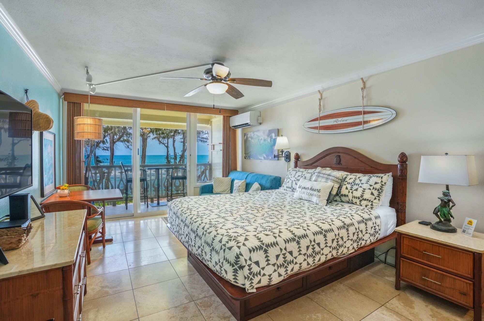 A hotel room with a bed, a desk, a chair, a ceiling fan, and a view of the ocean.