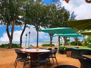 Ein Sitzbereich im Freien mit einem runden Tisch und Stühlen, umgeben von Bäumen und Sonnenschirmen, mit Blick auf Strand und Meer.