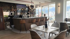 A modern bar area with a city view, a dining space with white tables and brown chairs, and a large window offering a panoramic view. - Zen Hotel Quito (Quito)