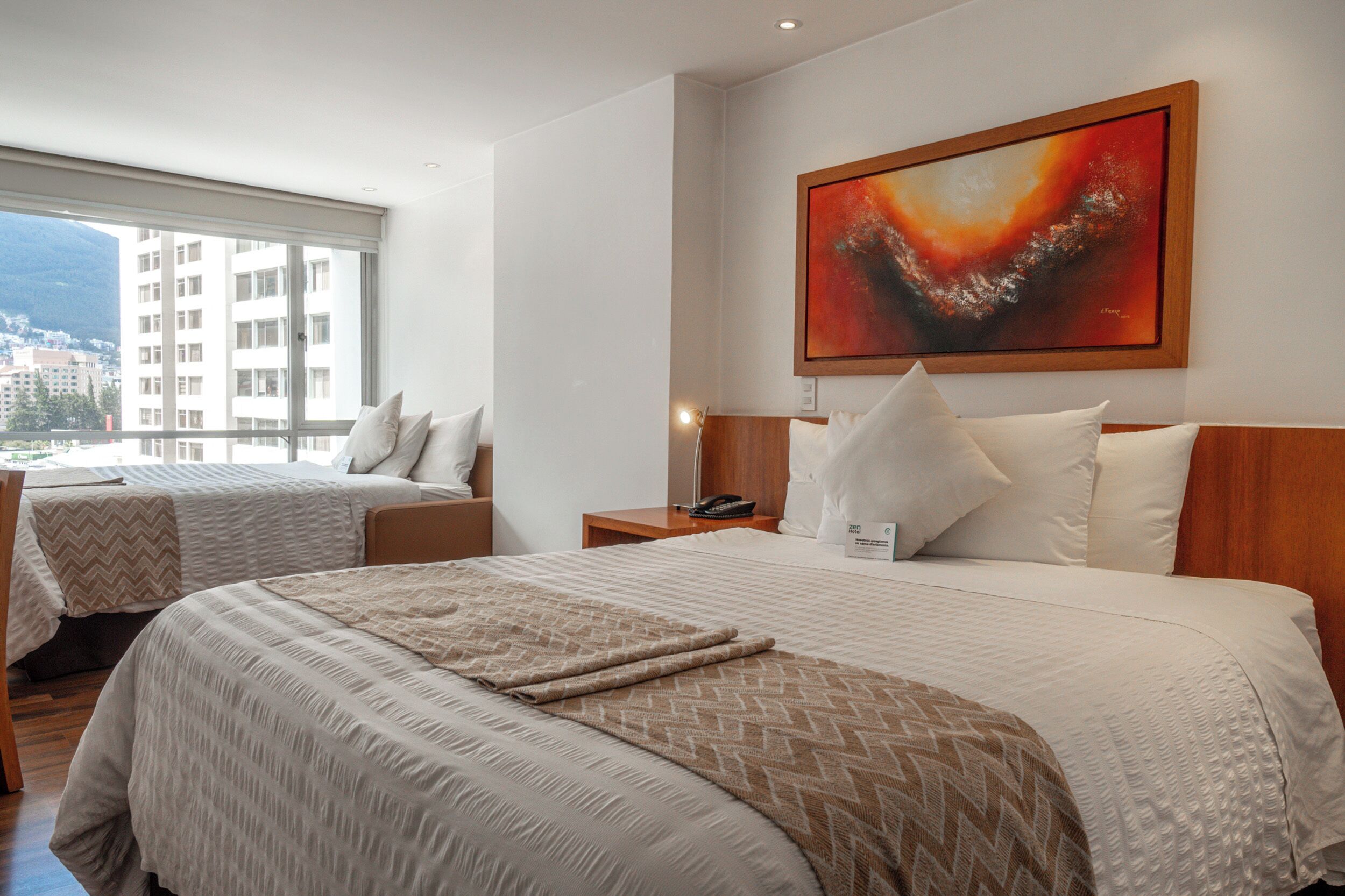Ein Hotelzimmer mit zwei Betten, einem großen Fenster, einem Gemälde an der Wand und Blick auf die Stadt.