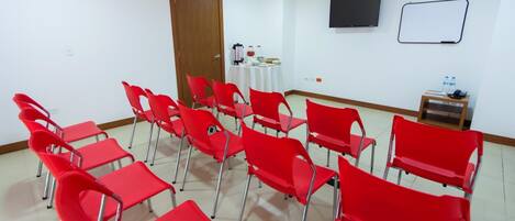 Un salón de conferencias con sillas rojas dispuestas en filas, una pizarra, un televisor y una pequeña mesa con botellas de agua y un teléfono.