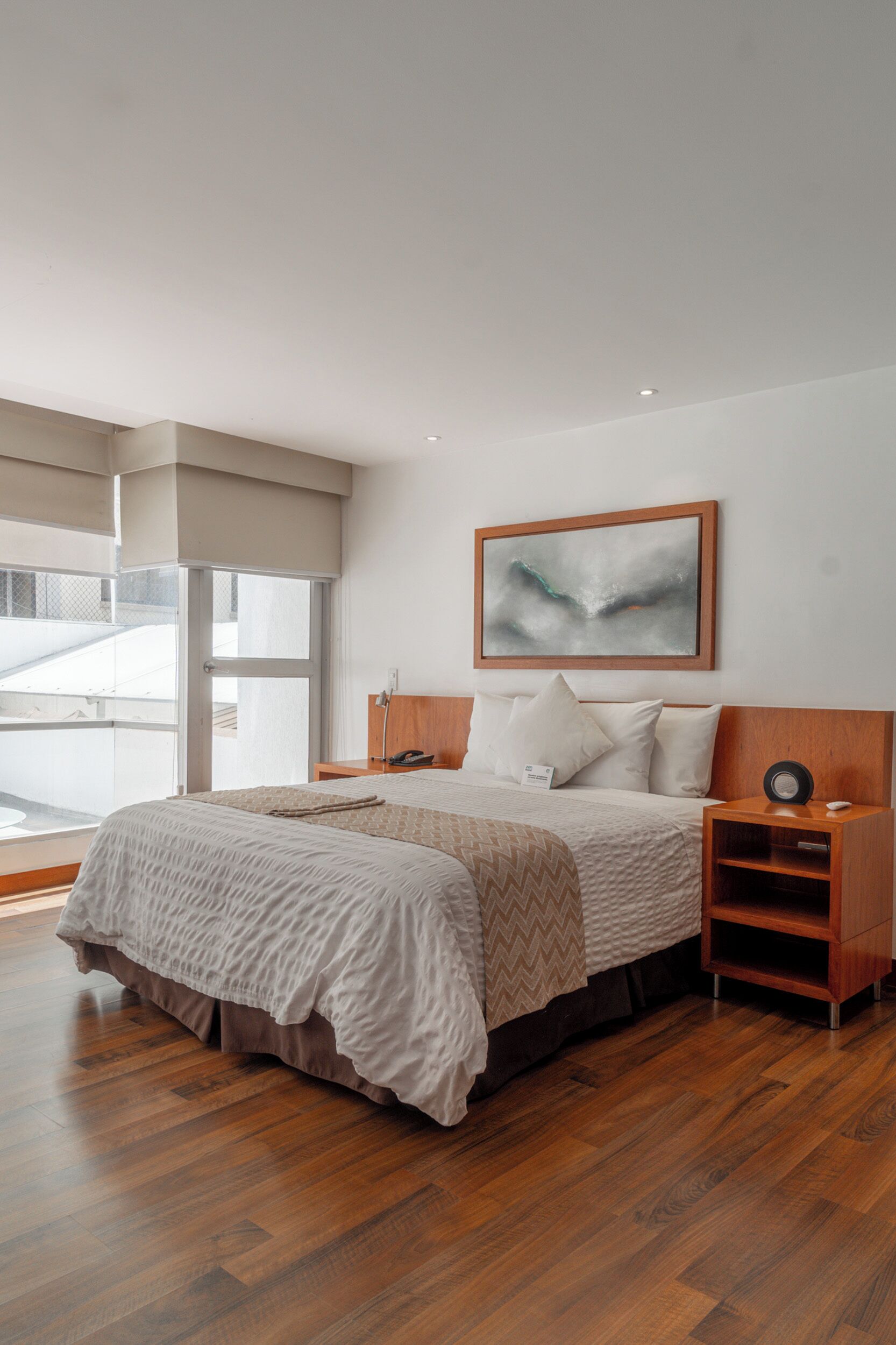 A modern bedroom with a large bed, wooden headboard, a bedside table with a lamp, and a large window with a view of the outside.