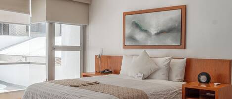 A modern bedroom with a large bed, wooden headboard, a bedside table with a lamp, and a large window with a view of the outside.