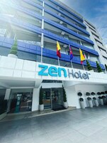 Un hotel moderno con un llamativo letrero que dice 'Zen Hotel', varias banderas y un área de entrada bien cuidada con plantas en macetas.