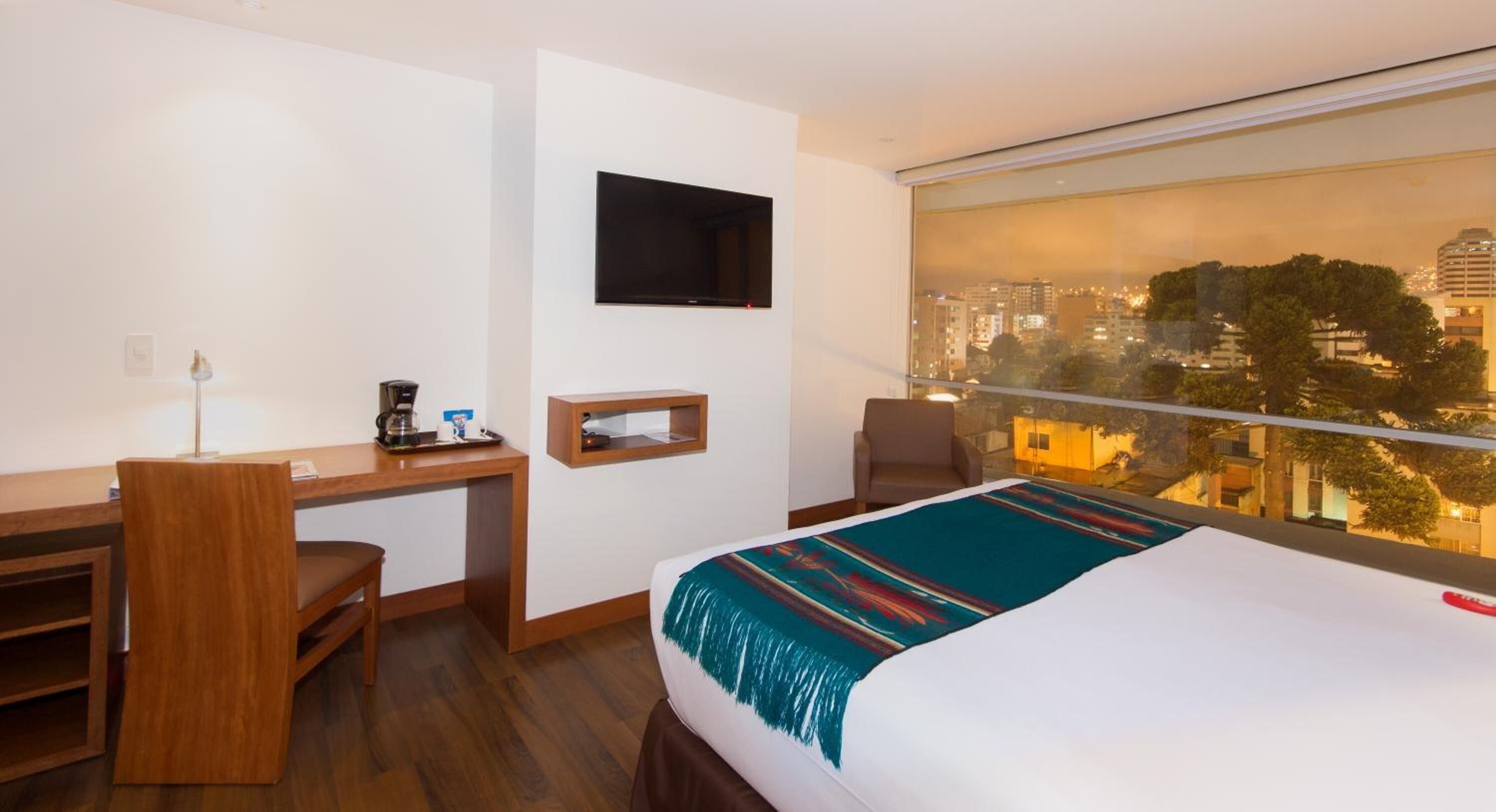 A hotel room with a bed, a desk, a chair, a TV, and a large window with a city view.