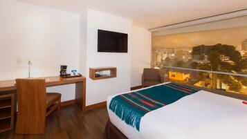 A hotel room with a bed, a desk, a chair, a TV, and a large window with a city view.