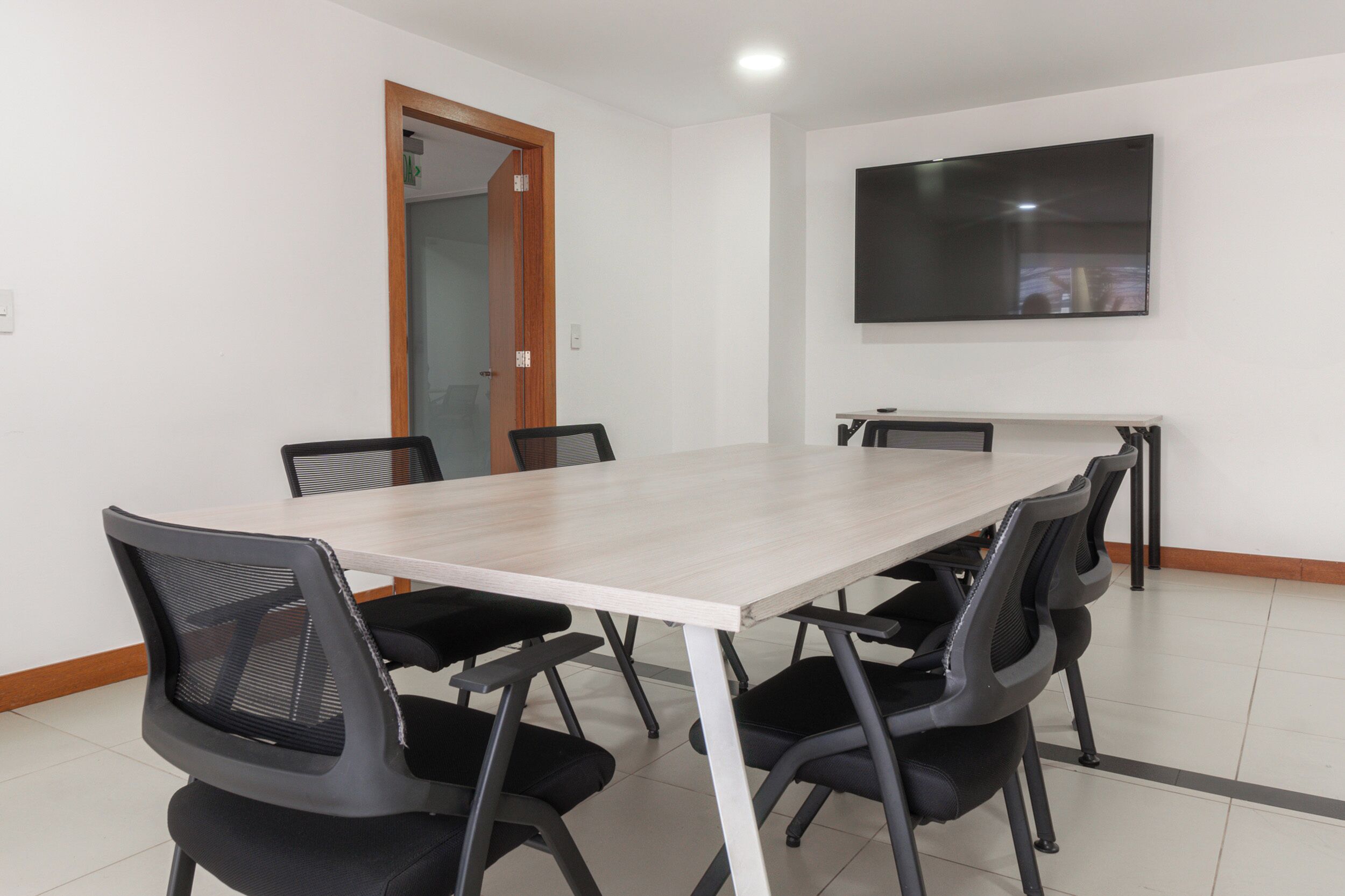 Una sala de conferencias con una mesa larga, seis sillas, un televisor de pantalla plana y una puerta que da a otra habitación.