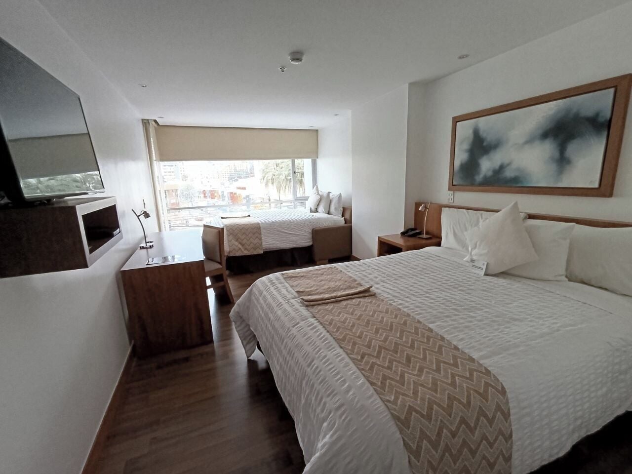 A hotel room with a large bed, a smaller bed, a TV, a desk, and a window with a view of buildings.