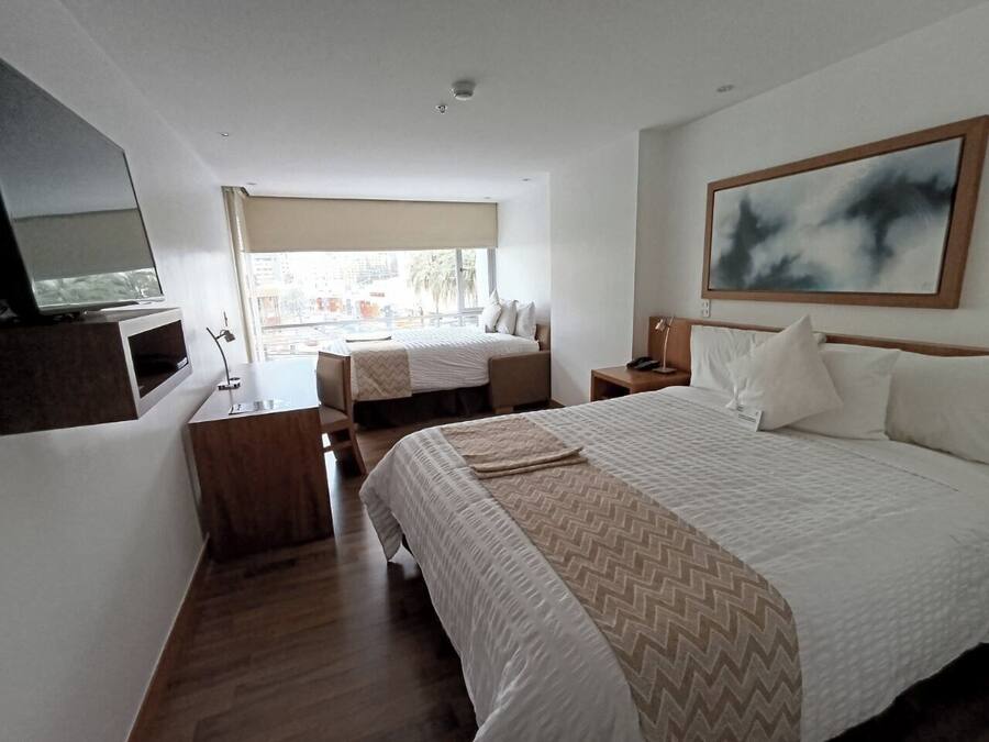 A hotel room with a large bed, a smaller bed, a TV, a desk, and a window with a view of buildings.
