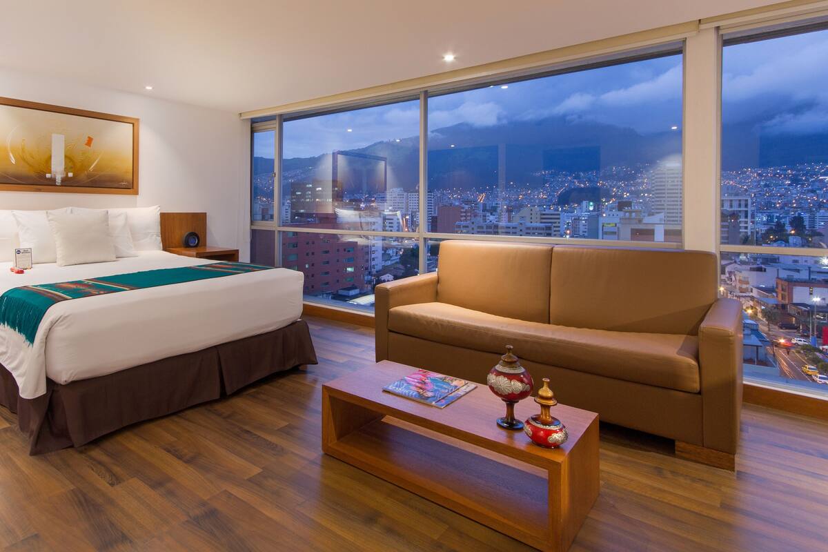 A hotel room with a large bed, a sofa, a coffee table, and a city view through large windows.