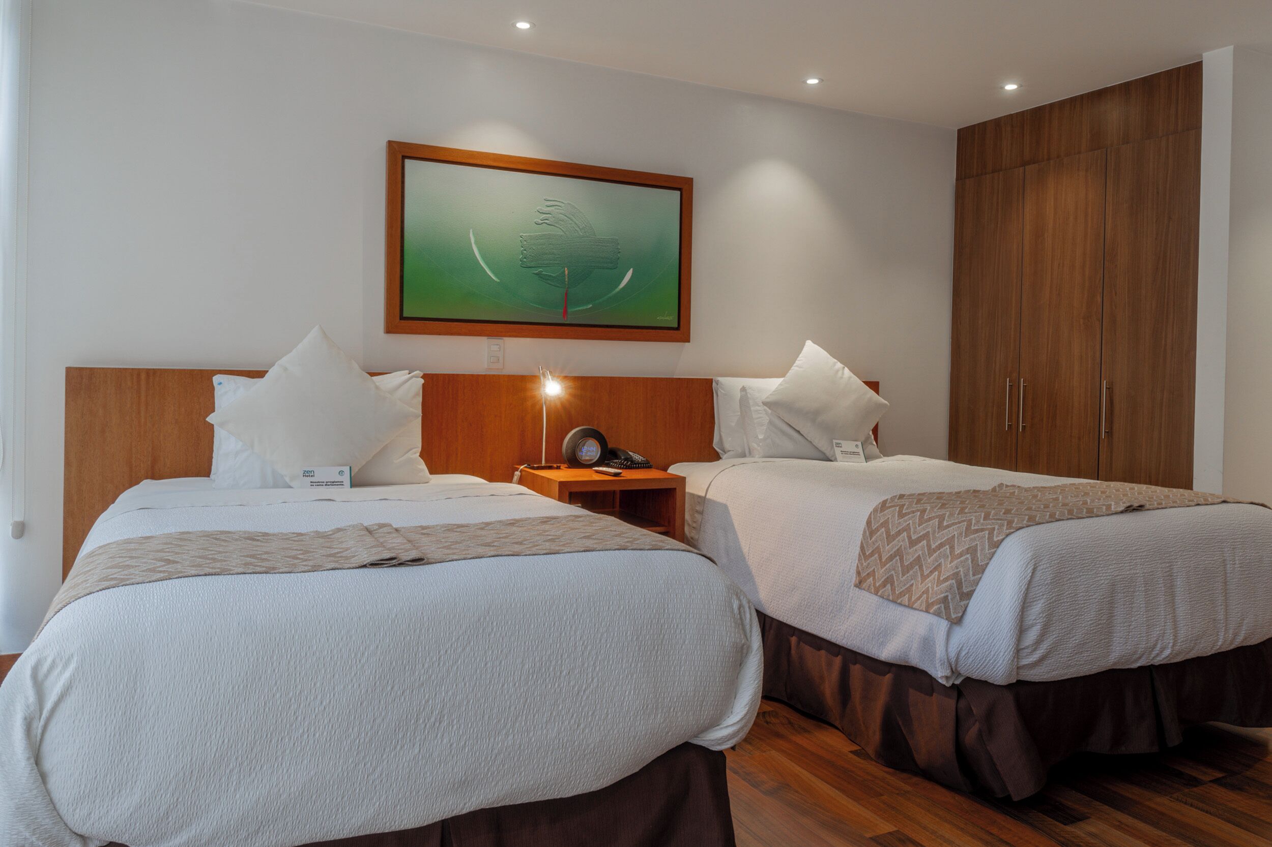 A hotel room with two beds, a nightstand with a lamp, a framed artwork on the wall, and a wardrobe.