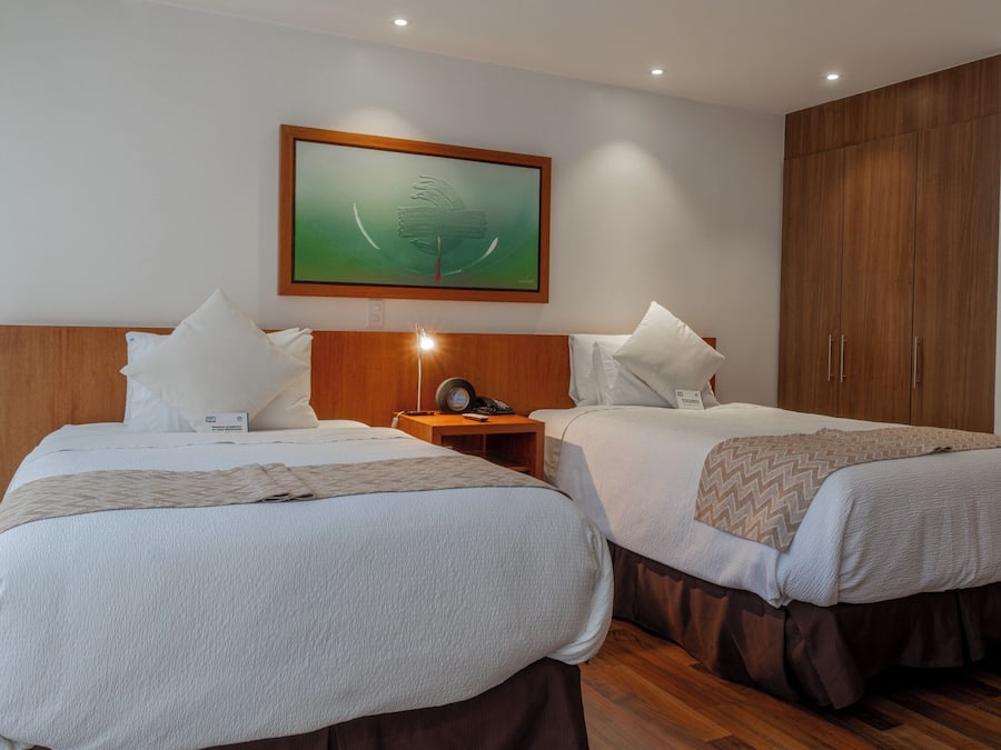 A hotel room with two beds, a nightstand with a lamp, a framed artwork on the wall, and a wardrobe.