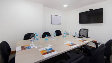 Un salón de conferencias con una larga mesa, sillas negras, botellas de agua, vasos y una pantalla de proyector.