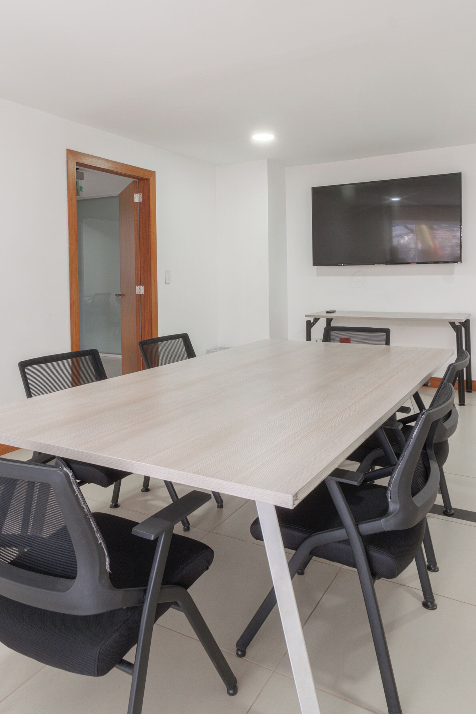 A conference room with a long table, six chairs, a TV on the wall, and a wooden door.