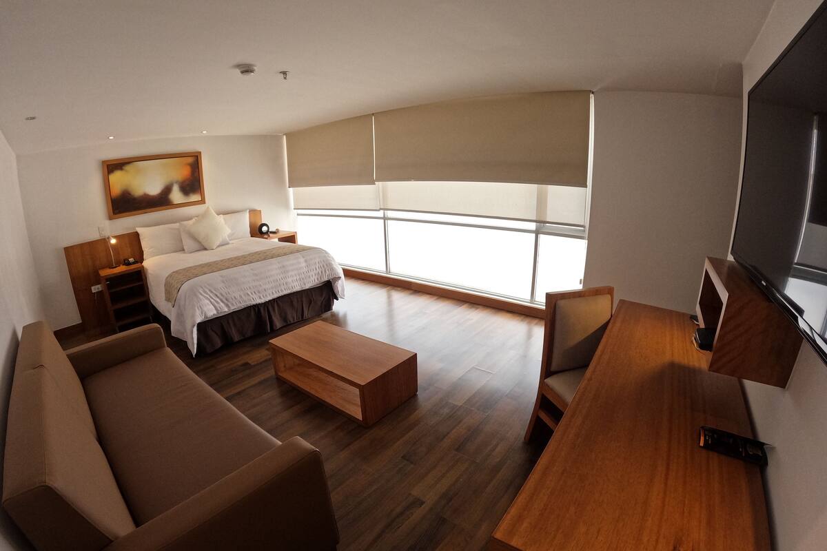 A modern hotel room with a bed, a sofa, a coffee table, a TV, and a large window with blinds.