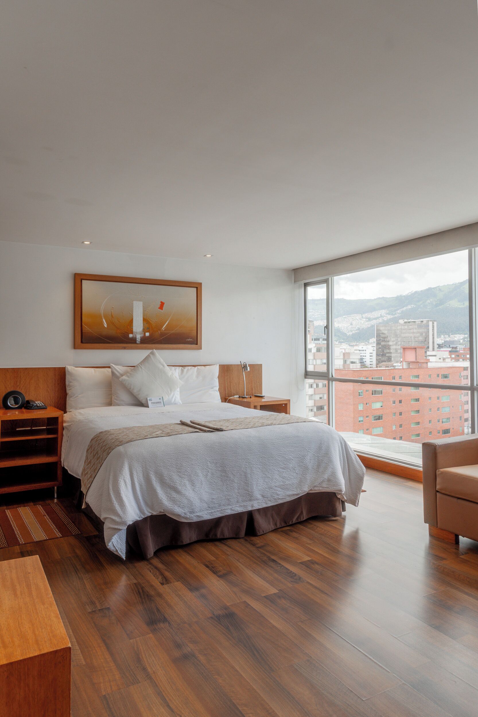 Una habitación de hotel moderna con una cama grande, piso de madera, vista a la ciudad y un cuadro en la pared.