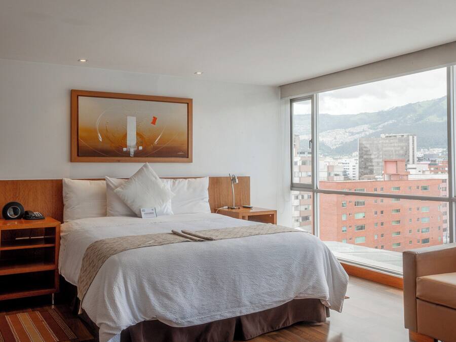 Una habitación de hotel moderna con una cama grande, piso de madera, vista a la ciudad y un cuadro en la pared.