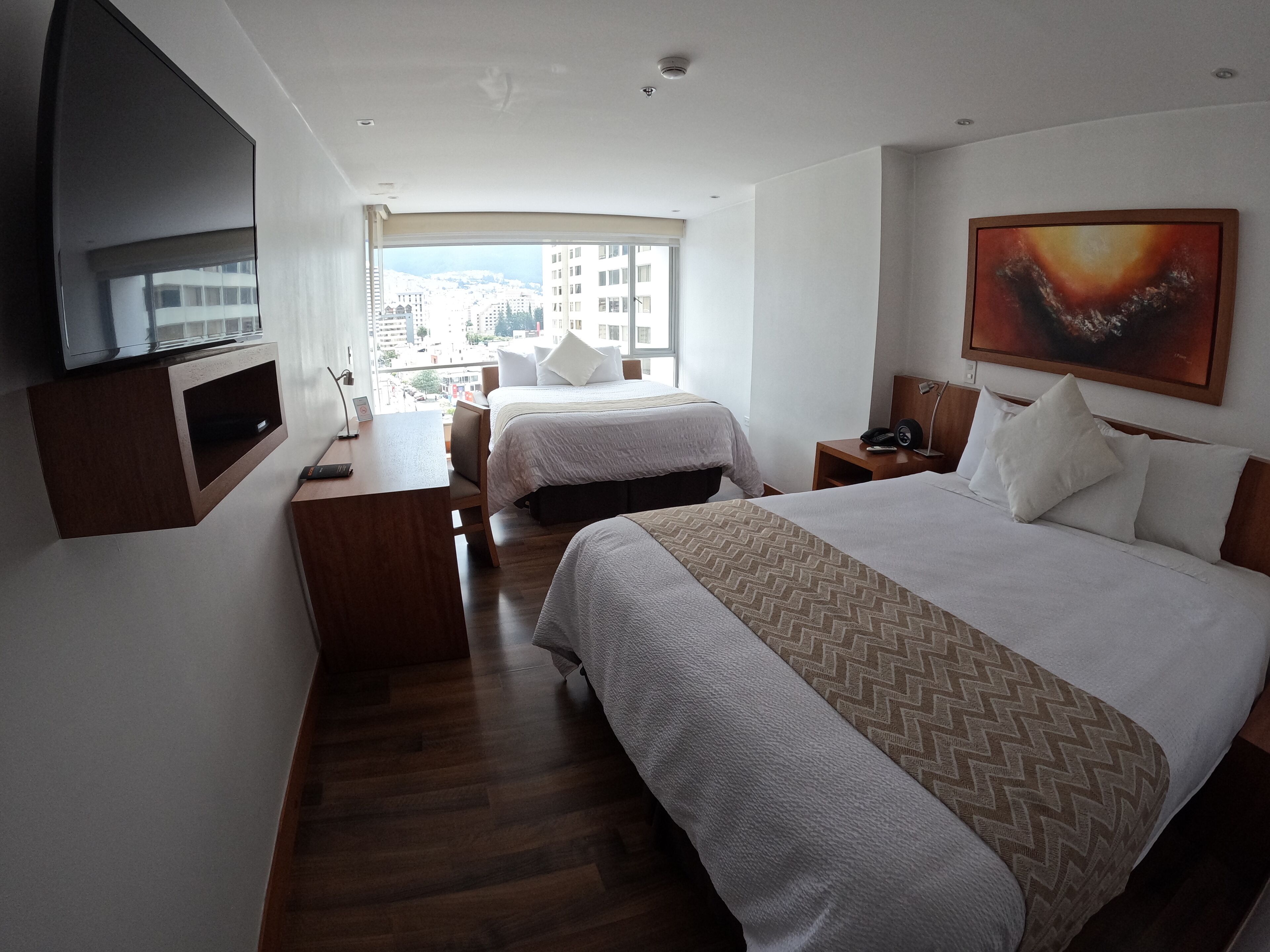 A hotel room with two beds, a desk, a TV, and a large window with a city view.