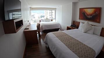 A hotel room with two beds, a desk, a TV, and a large window with a city view.