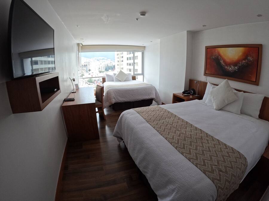 A hotel room with two beds, a desk, a TV, and a large window with a city view.