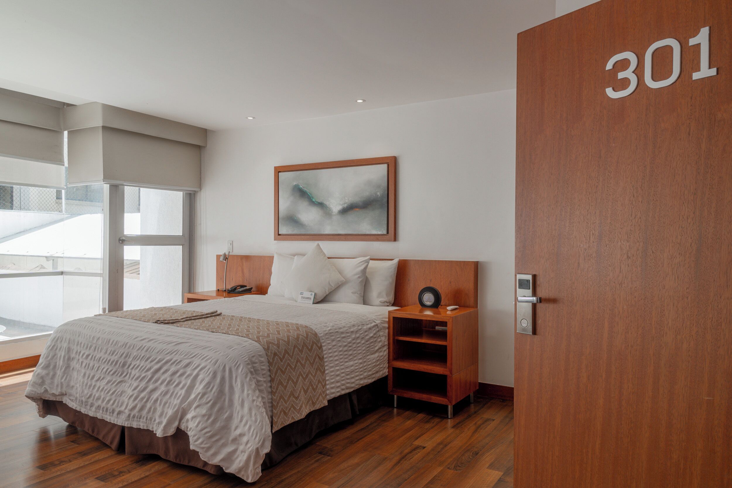 A modern hotel room with a large bed, a bedside table with a phone, a wall-mounted painting, and a door with the number 301.