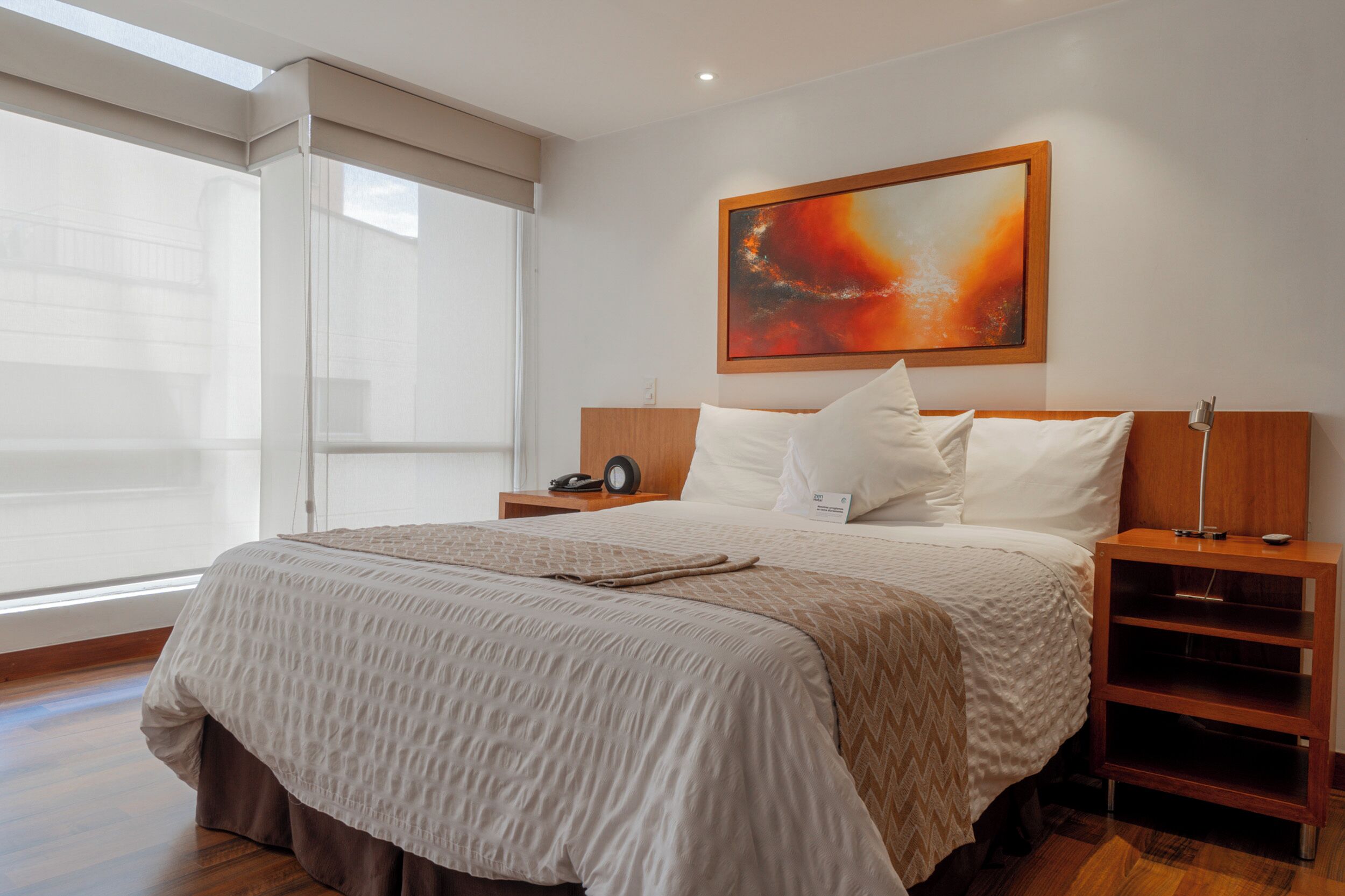 A hotel room with a large bed, a bedside table with a lamp, a painting on the wall, and a window with blinds.