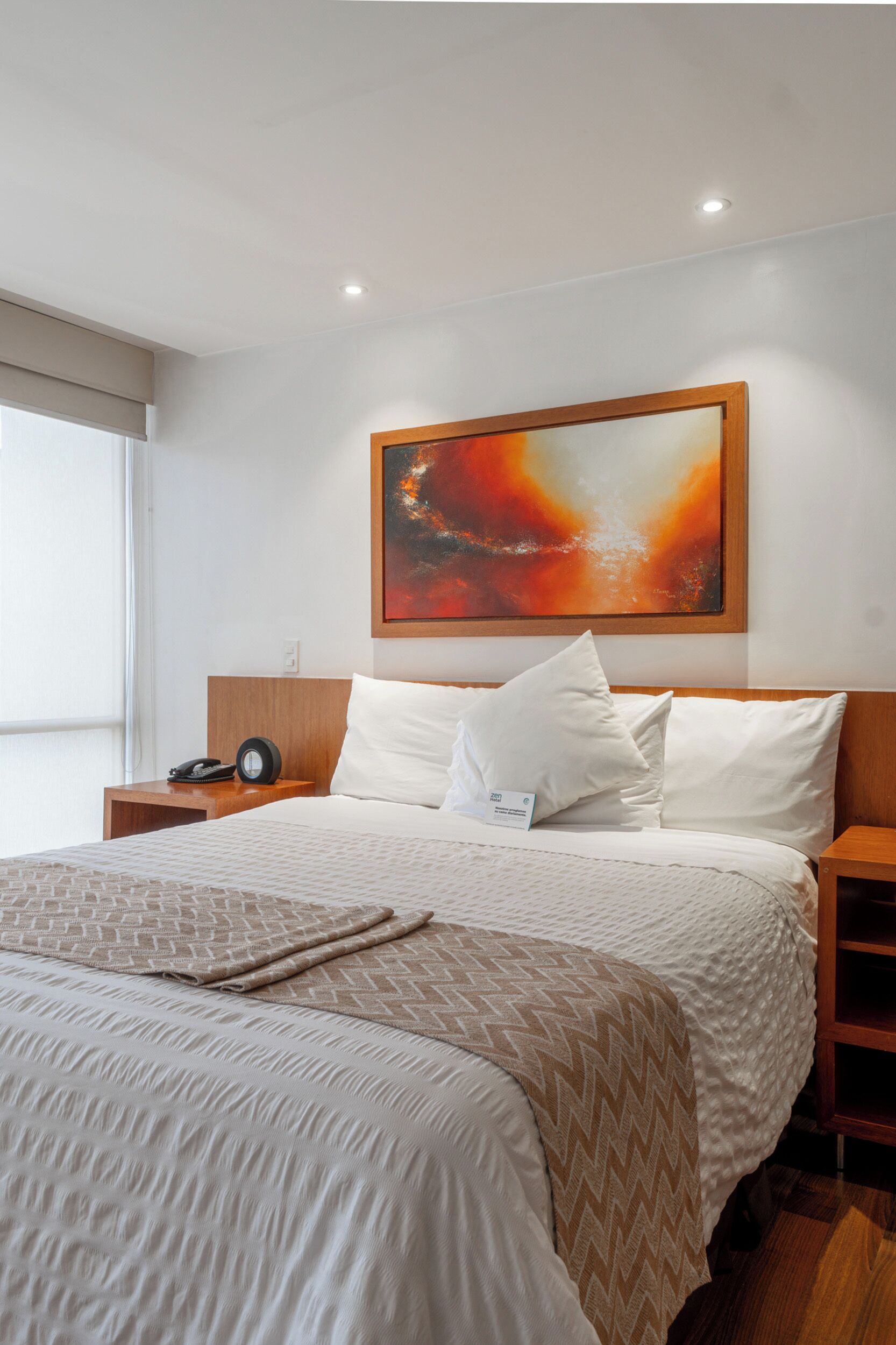 A hotel room with a large bed, a nightstand, a wall-mounted painting, and a window with blinds.