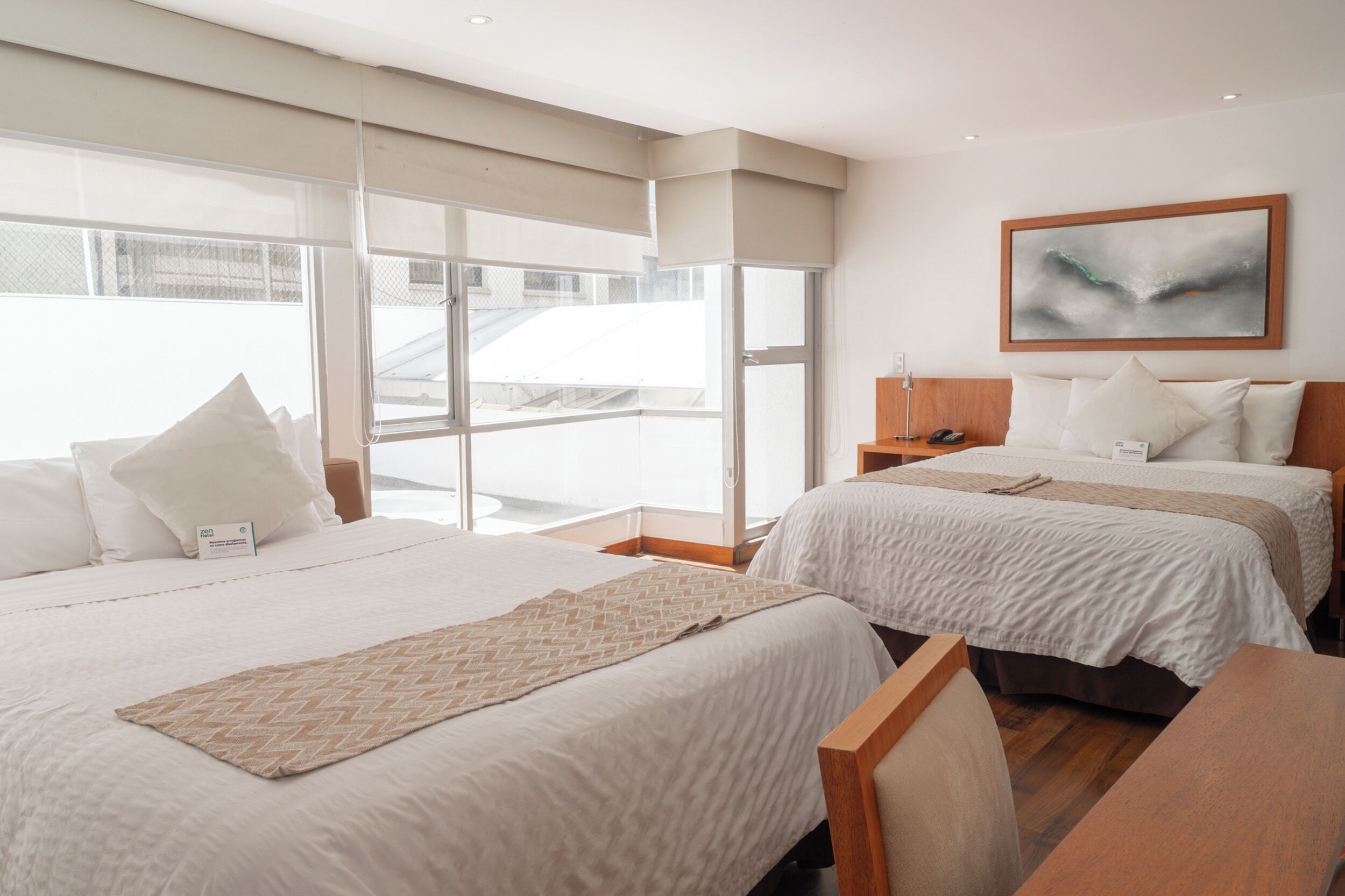 A hotel room with two beds, a wooden desk, a large window with a view of a building, and a painting on the wall.
