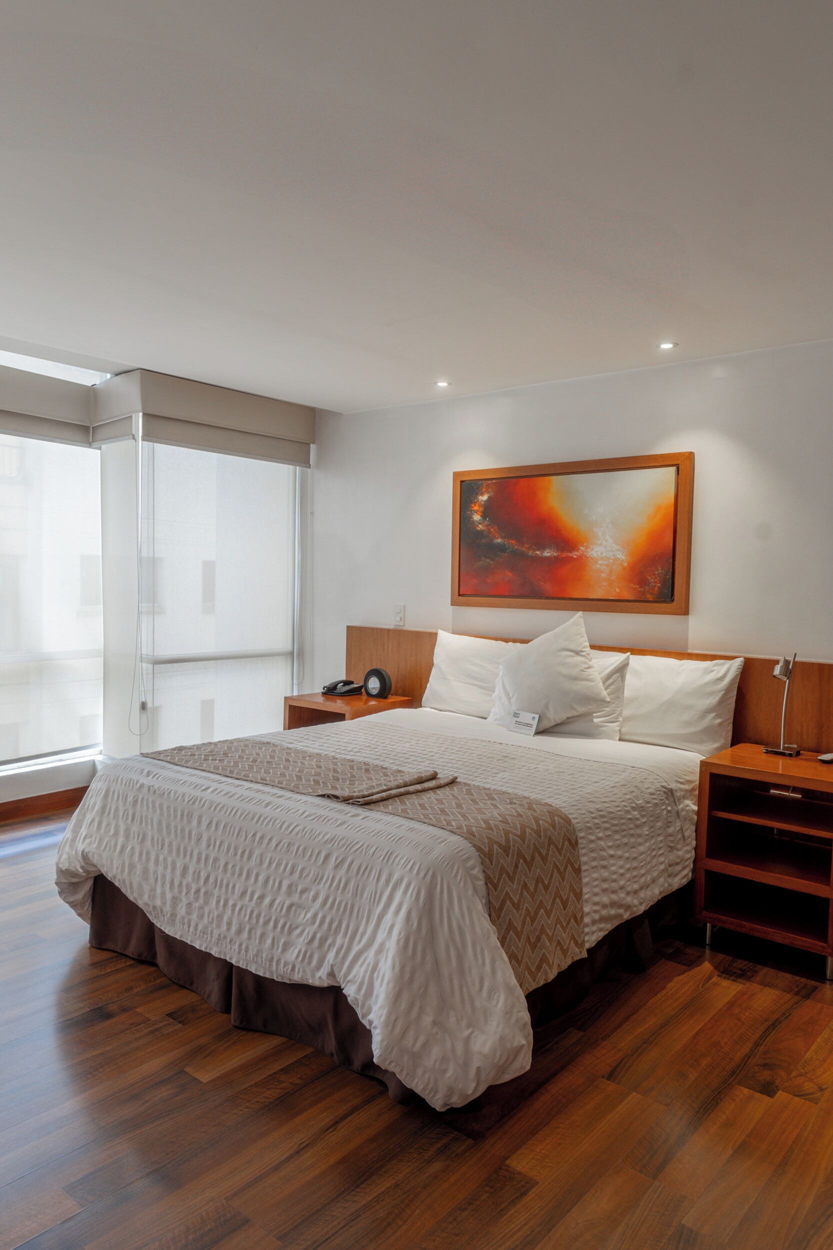 A modern bedroom with a large bed, wooden flooring, a glass door leading to a balcony, and a painting on the wall.