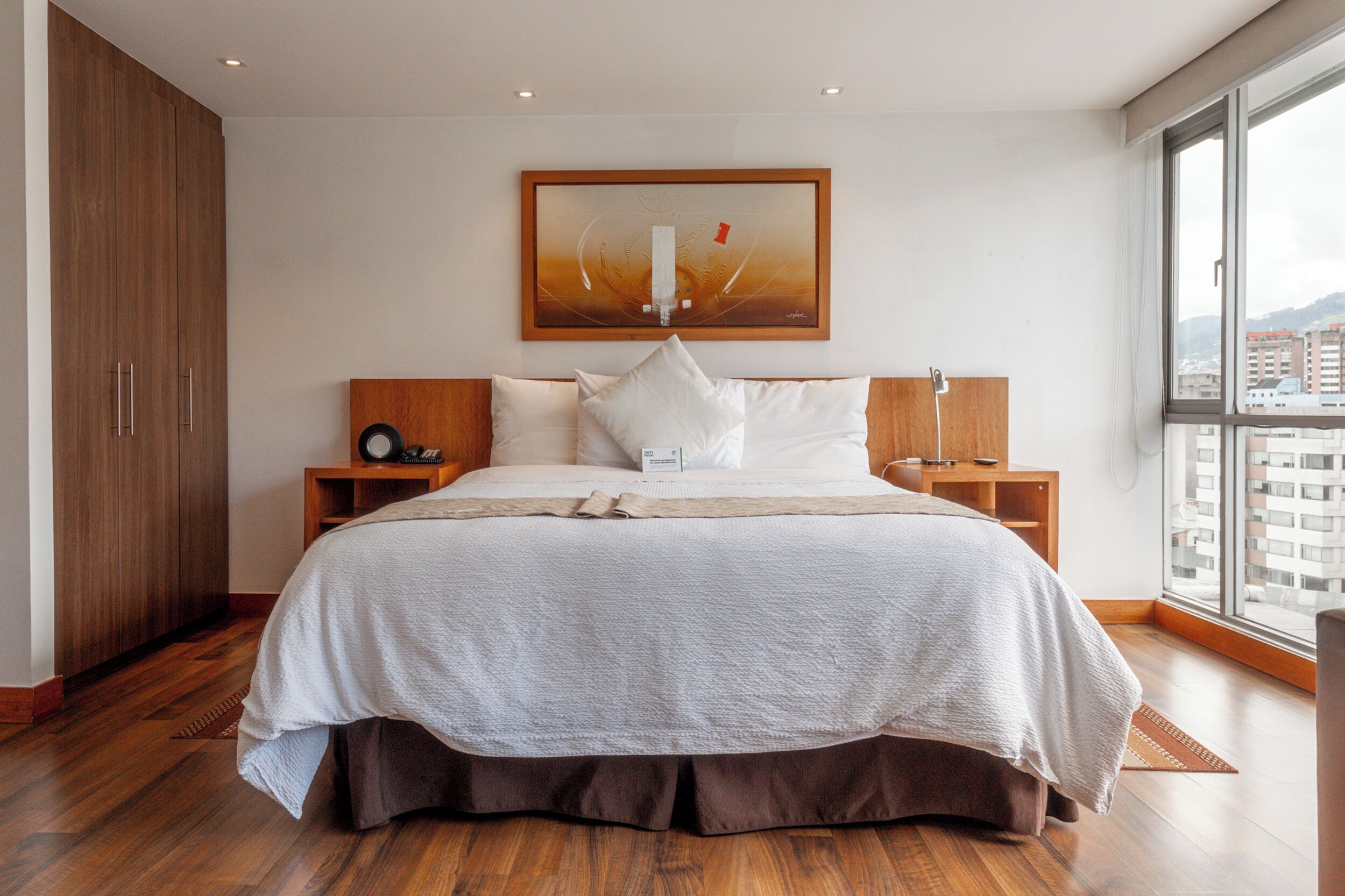 A modern bedroom with a large bed, wooden wardrobe, bedside tables, and a city view through the window.