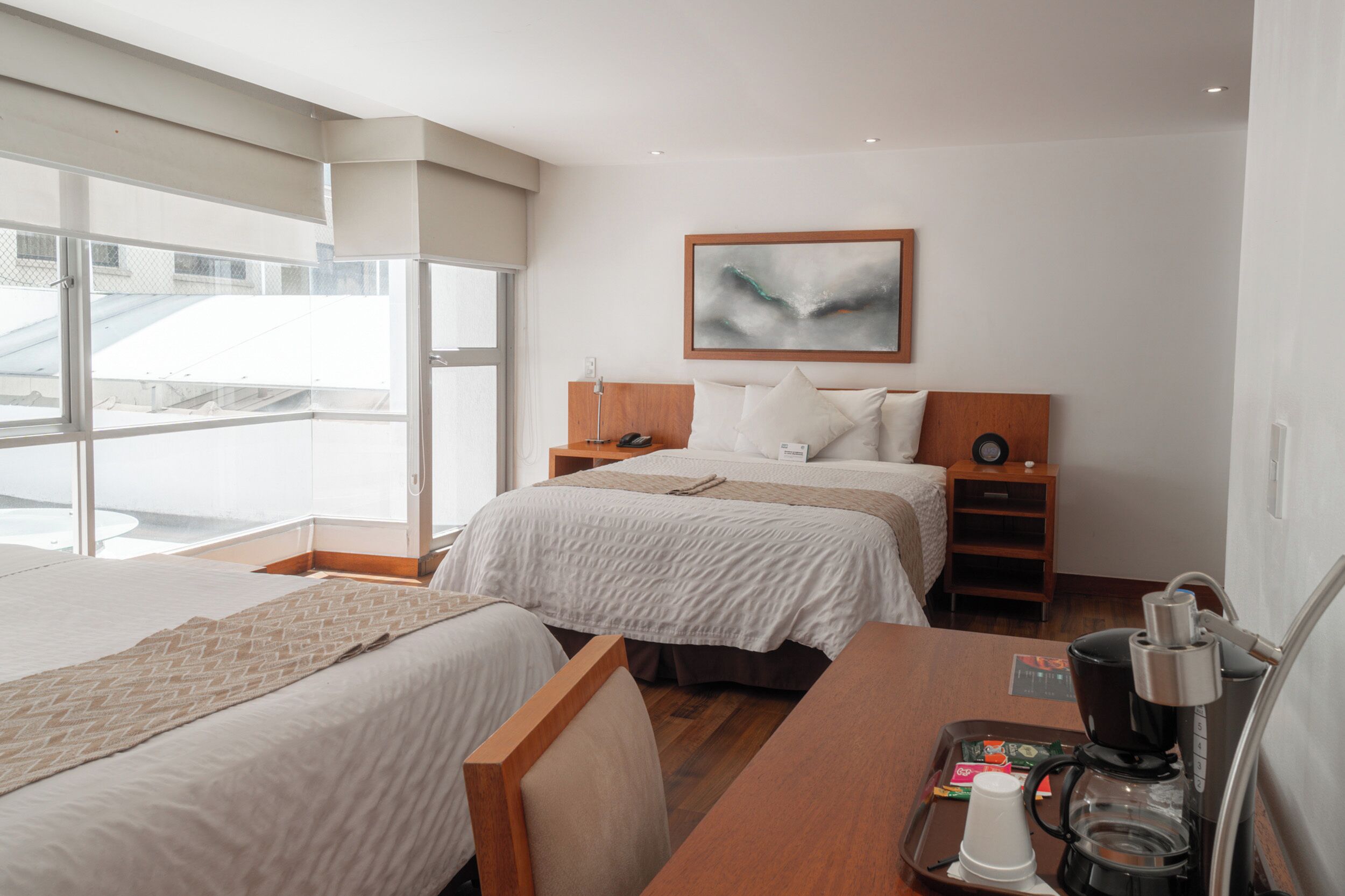 Quarto de hotel com duas camas, uma escrivaninha de madeira, máquina de café e uma janela grande com vista para a cidade.