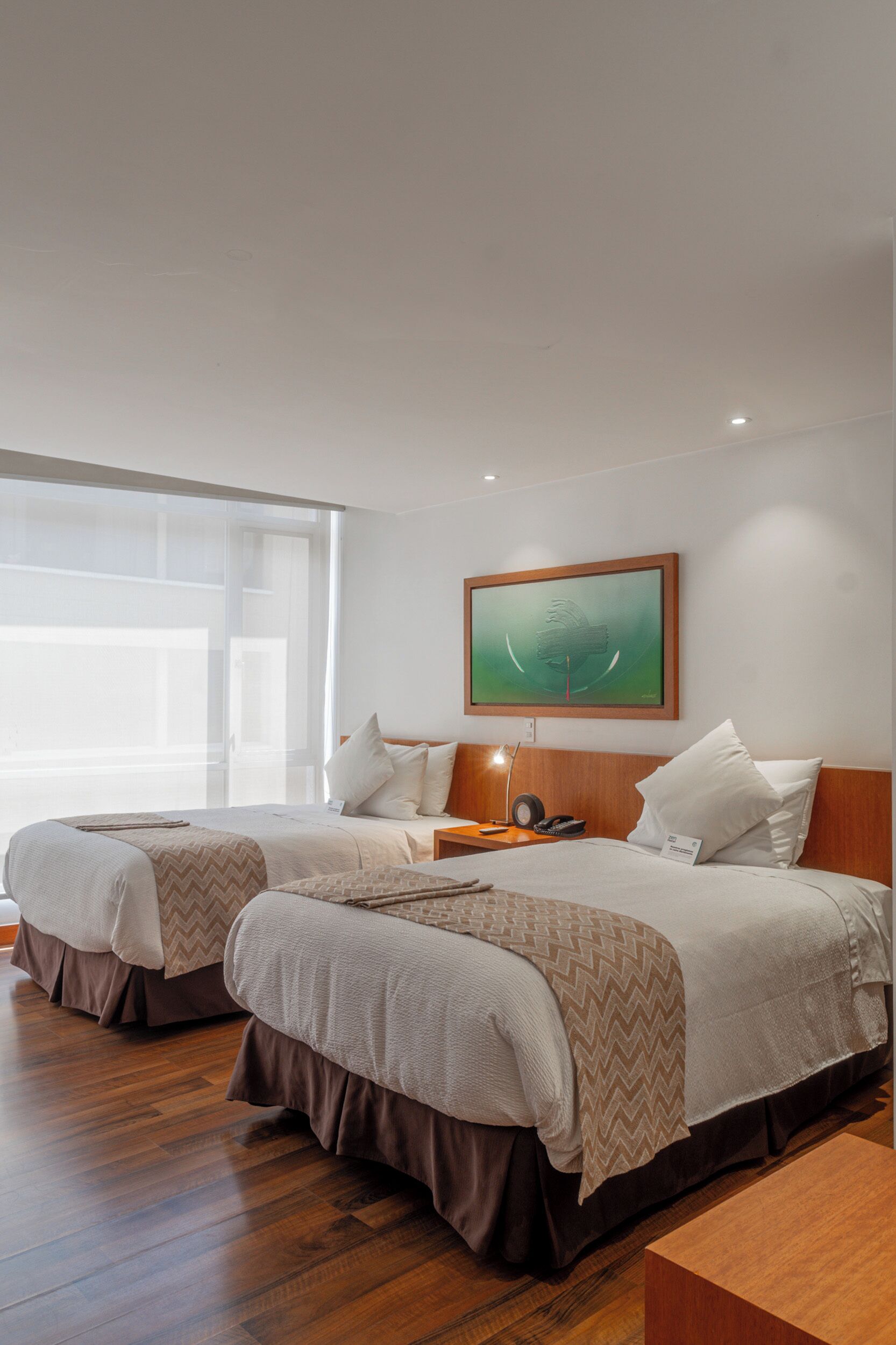 A hotel room with two beds, a large window, a painting on the wall, and a nightstand with a lamp.
