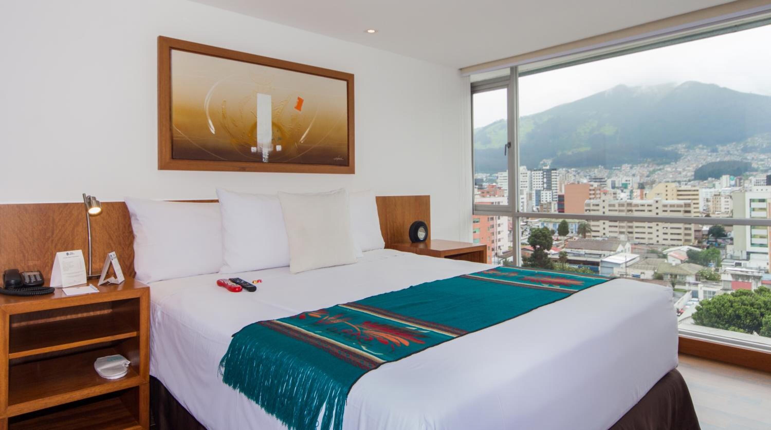 A hotel room with a large bed, a nightstand with a lamp, a clock, and a view of a cityscape and mountains through the window.