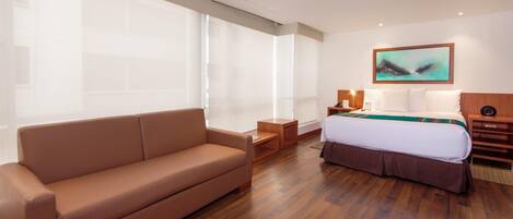 A hotel room with a large window, a bed, a sofa, a nightstand, and a painting on the wall.