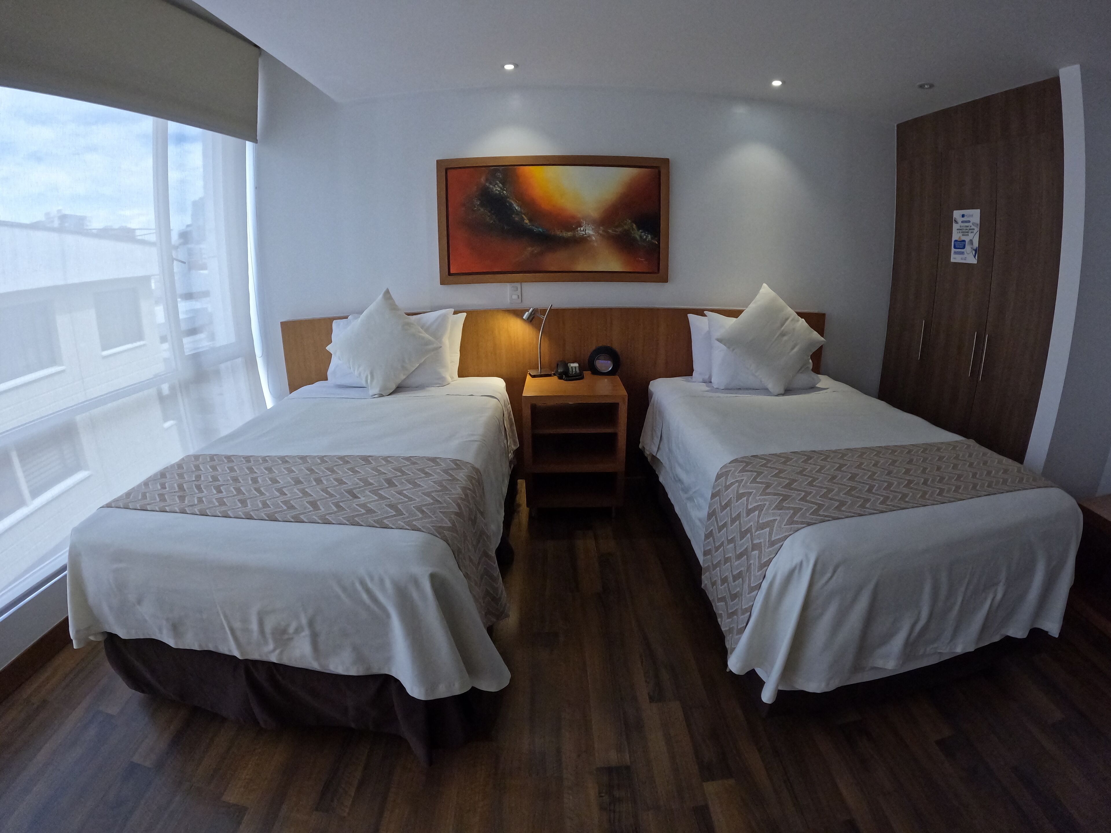 A hotel room with two beds, a wooden wardrobe, a painting on the wall, and a window with a view of buildings.