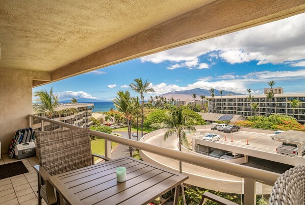 A balcony with a view of a parking area, palm trees, and a building with a pool.