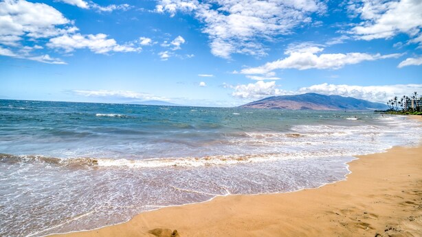Una playa con agua azul cristalina, orilla arenosa y montañas a lo lejos.