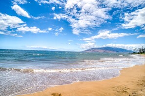 A beach with clear blue water, sandy shore, and distant mountains.