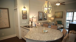 A kitchen with a central island, white cabinetry, and a dining area with wicker chairs. - Luxury Condo at Village Pier, King Bed,Full Kitchen, Polaris Ranger for Rent (St. Simons Island)