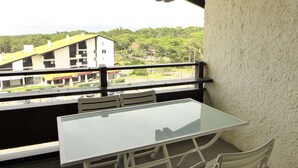 A balcony with a glass table and white chairs, overlooking a building with a green roof and trees.