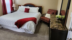 Quarto de hotel com uma cama, uma cadeira, uma escrivaninha com espelho e uma luminária.