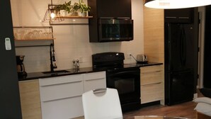 Cozinha moderna com micro-ondas, forno e geladeira pretos, uma mesa de jantar de vidro com cadeiras brancas e uma luminária pendente acima.