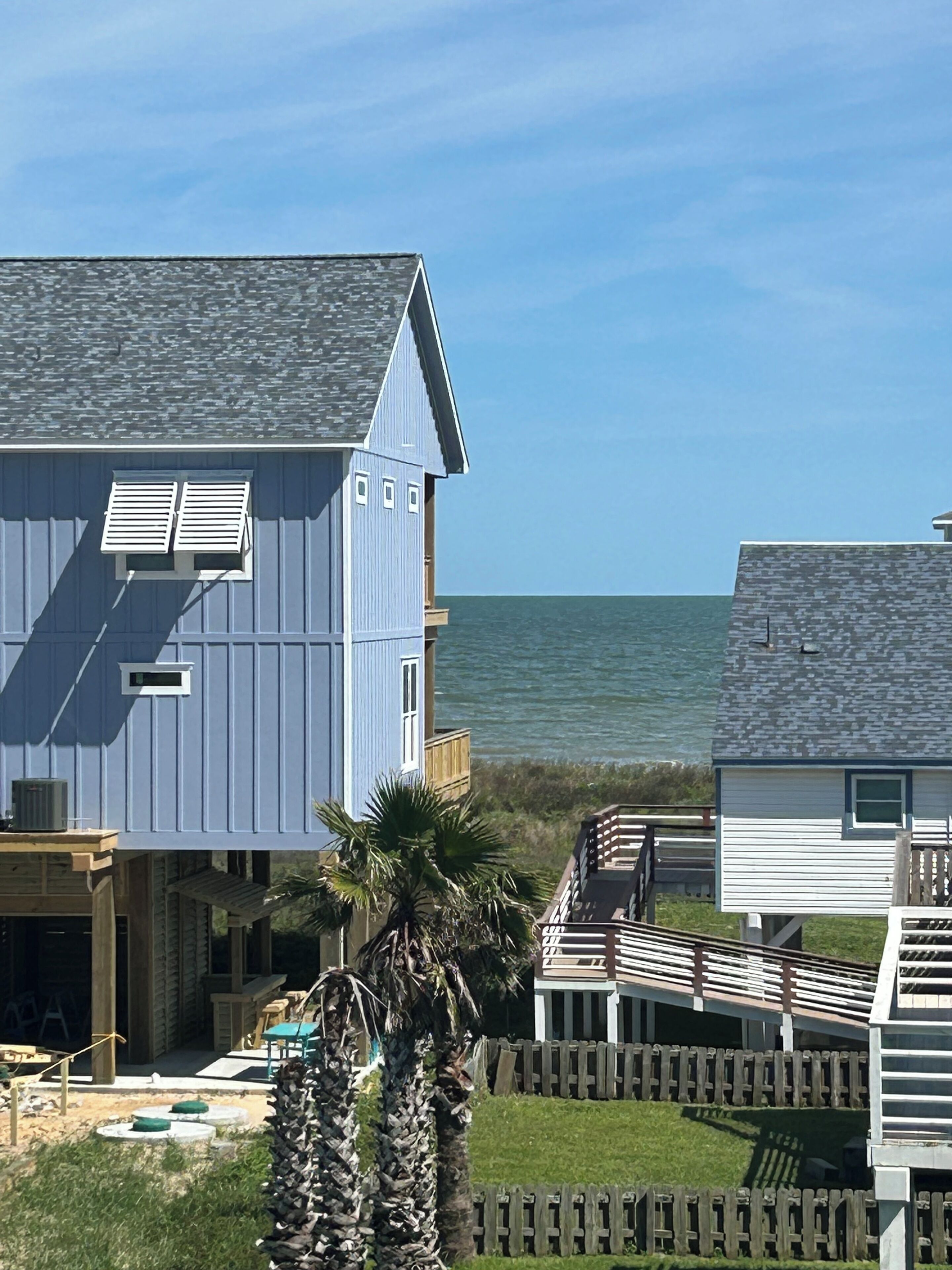 A coastal house with a balcony overlooking the sea, a wooden deck, and a fenced yard.