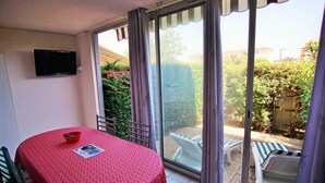 A room with a red patterned table, a television mounted on the wall, and a sliding glass door leading to an outdoor area with a garden and a wooden gate. - Residence with swimming pool and private parking space! (LA SEYNE SUR MER)