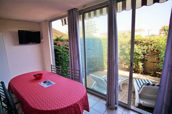 Una habitación con una mesa de diseño rojo, un televisor fijado a la pared y un ventanal corredero que da a un espacio exterior con jardín y una puerta de madera.