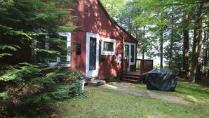 A red cabin with a porch, surrounded by dense forest. - Waterfront Cottage on Scenic Sportsman Pond (Fitzwilliam)