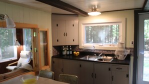 A kitchen with wooden cabinets, a window, and a dining area with a table and chairs. - Waterfront Cottage on Scenic Sportsman Pond (Fitzwilliam)