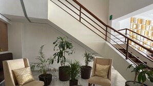 Um hall com piso de mármore, duas poltronas bege, uma mesa de madeira redonda e algumas plantas em vasos. Uma escada com corrimão leva a um andar superior.