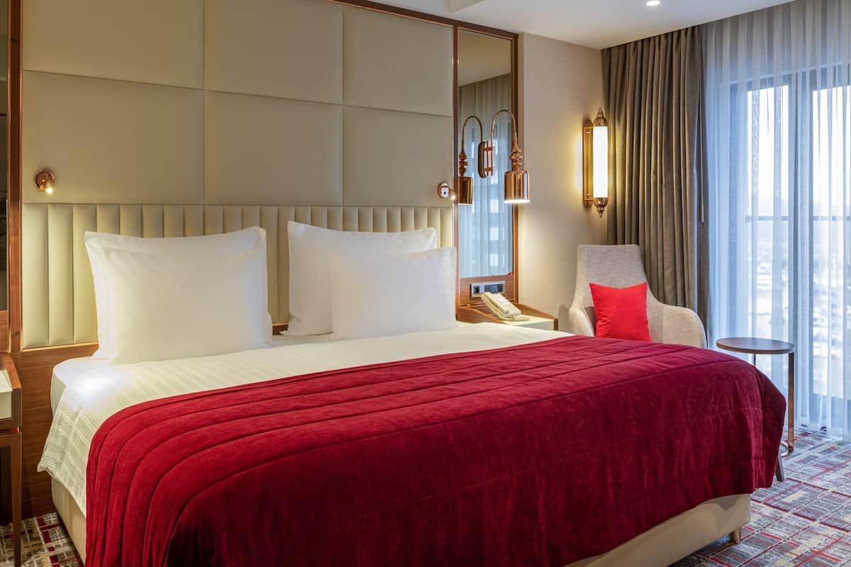 A hotel room with a large bed, a chair, a bedside table, a mirror, and a window with curtains.
